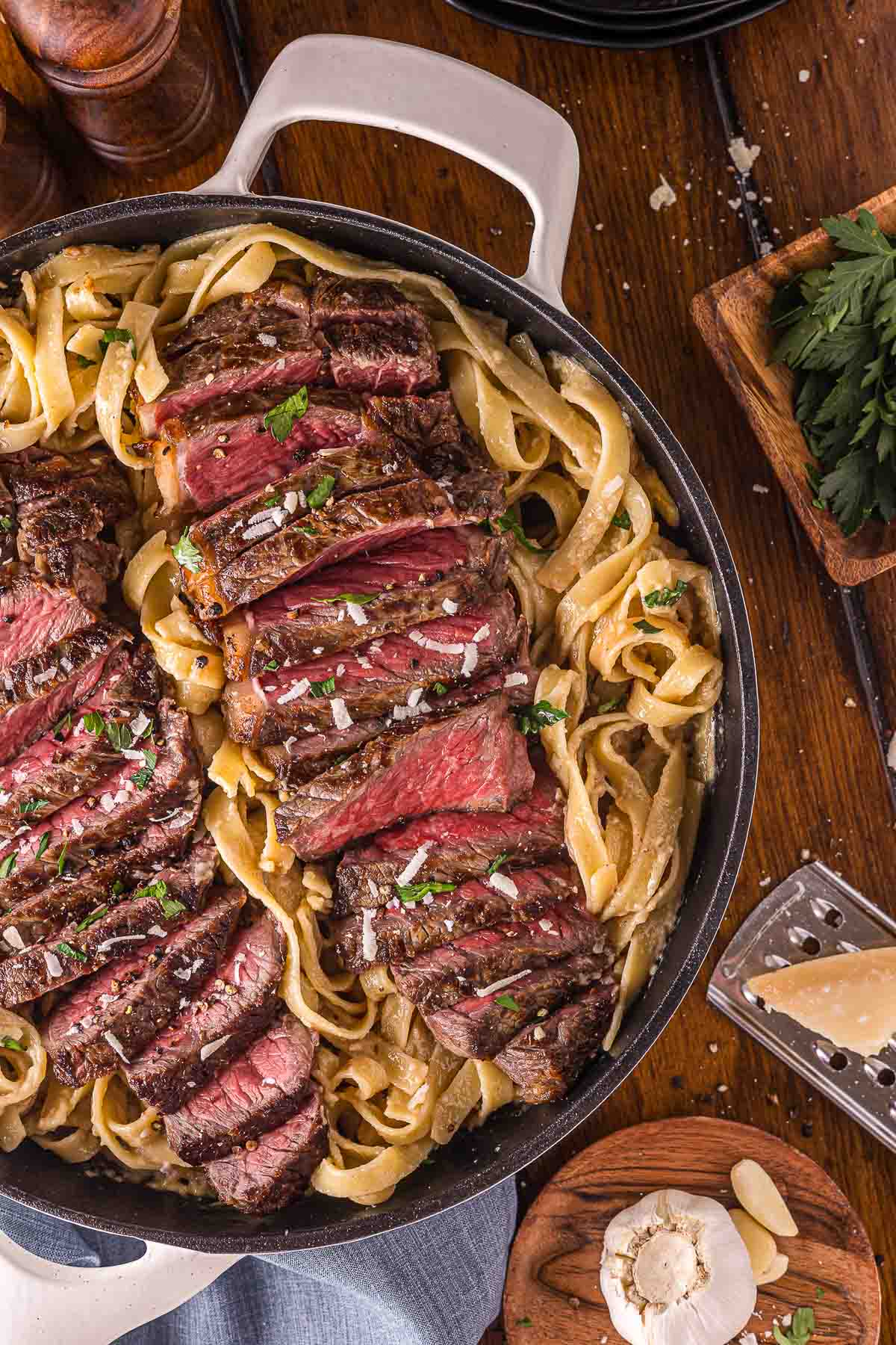 A skillet filled with sliced medium-rare steak arranged over fettuccine pasta, garnished with herbs and coarse salt. Nearby are a block of parmesan, garlic, and fresh parsley.