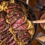 A skillet filled with sliced medium-rare steak Alfredo, garnished with parsley, on a wooden table with utensils and ingredients nearby.