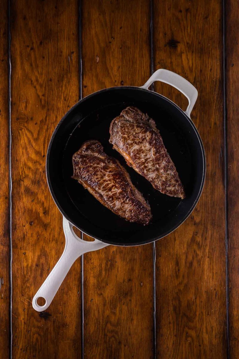Two cooked steaks in a white-handled black skillet placed on a wooden surface.