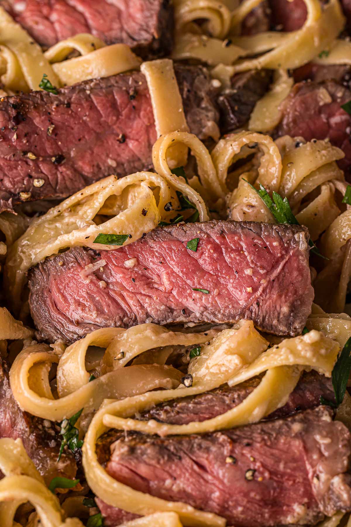 Close-up of medium-rare steak slices served on fettuccine pasta with a creamy Alfredo sauce and garnished with herbs.