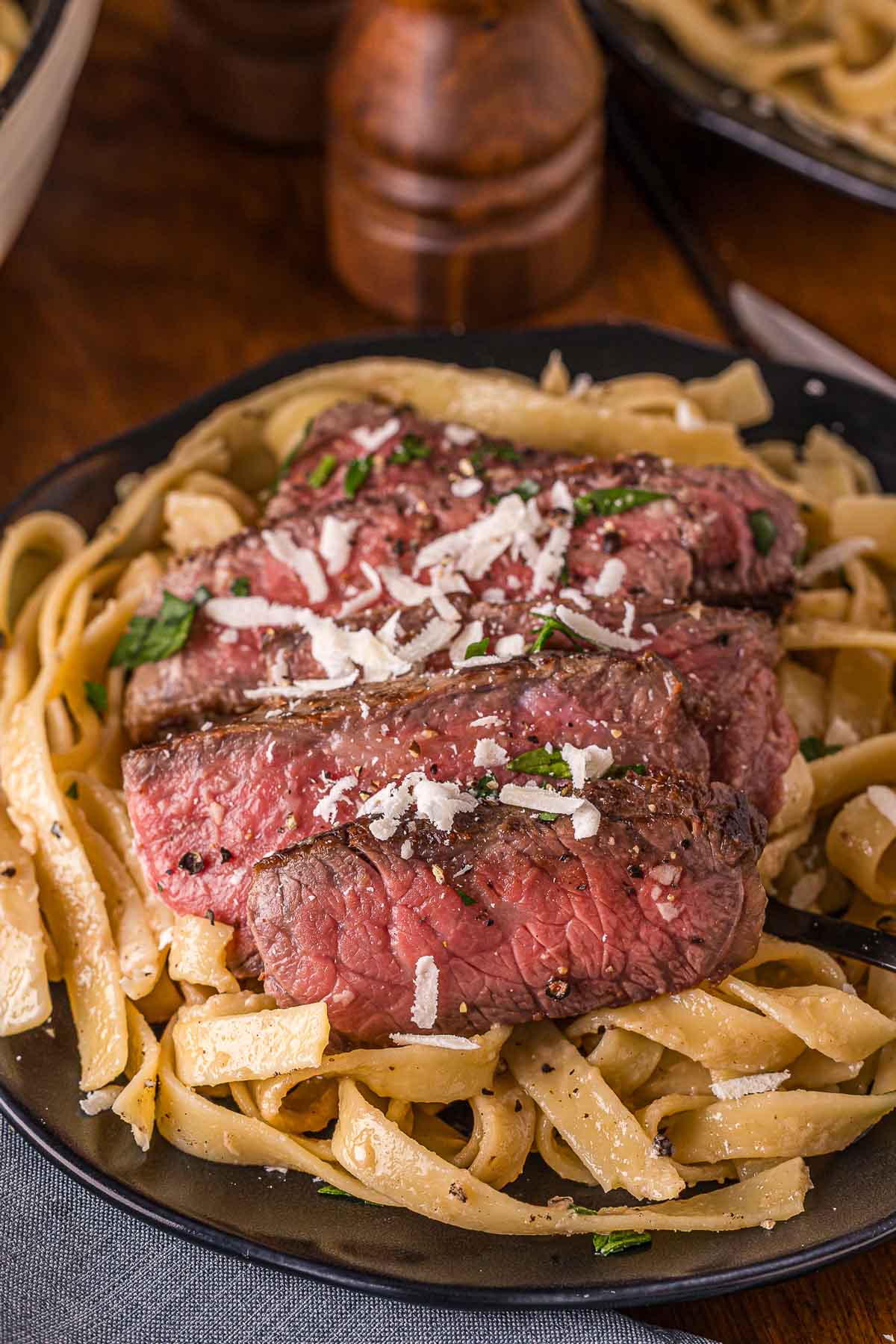 A plate of fettuccine Alfredo pasta topped with slices of medium-rare steak and sprinkled with grated cheese.