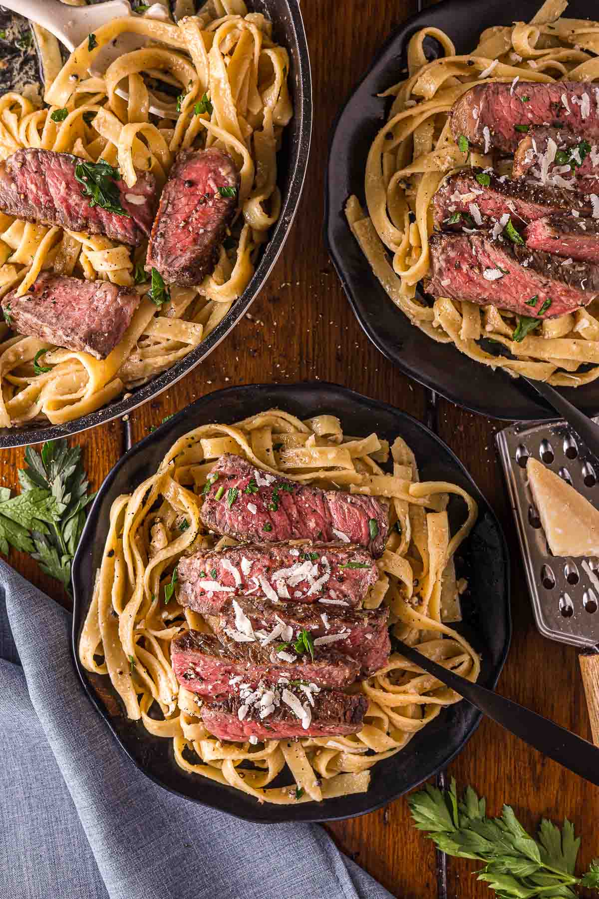 Three plates of steak alfredo with fettuccine pasta topped with sliced steak and grated cheese, garnished with herbs, on a wooden table with parsley and a cheese grater nearby.