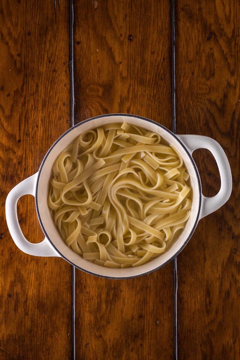 A white pot filled with cooked fettuccine pasta sits on a wooden table.