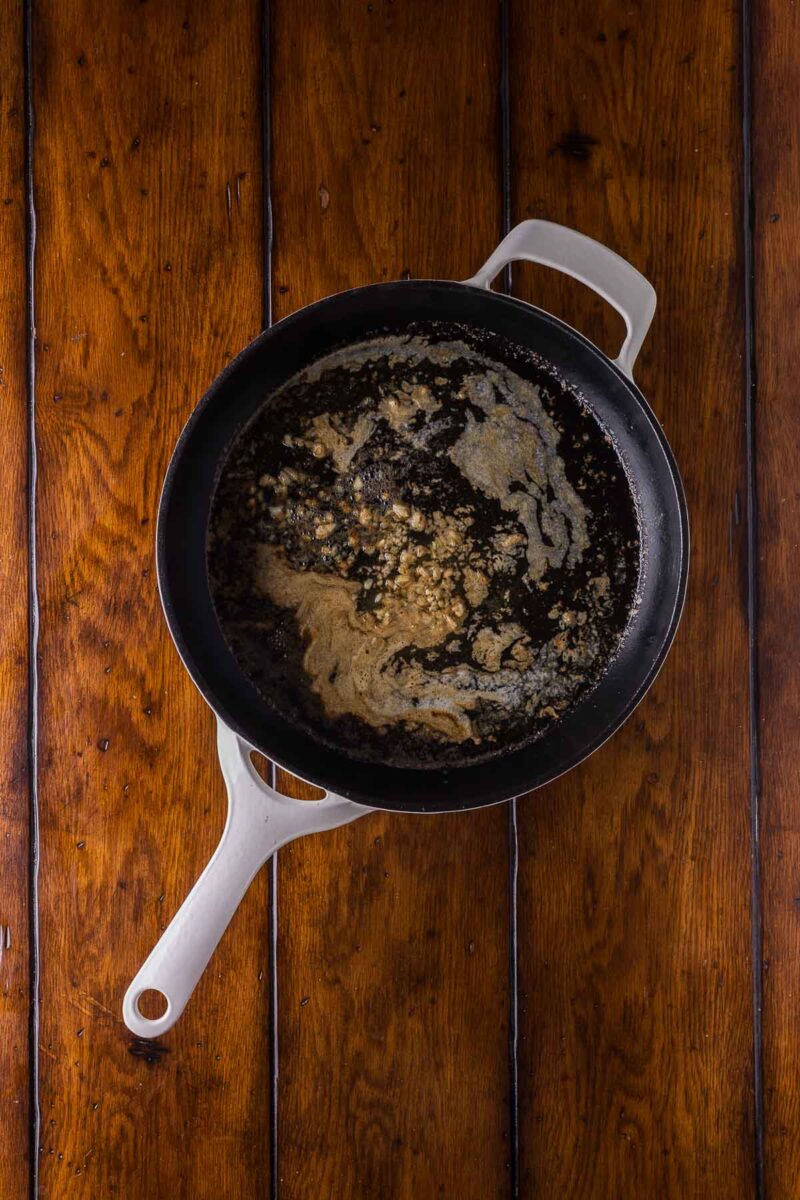 A white-handled skillet with browned butter and small bits of garlic sits on a wooden table.
