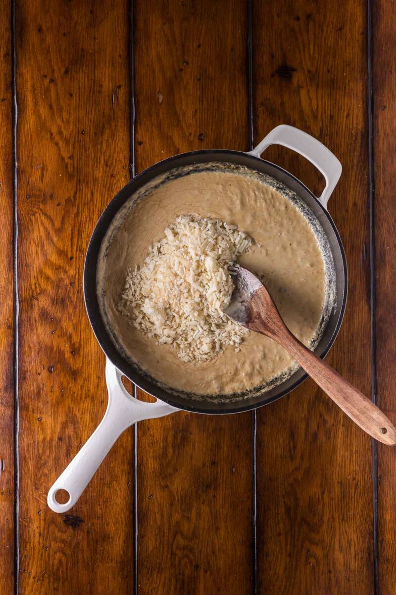 A white-handled pot with creamy sauce and cheese being stirred with a wooden spoon sits on a wooden table.