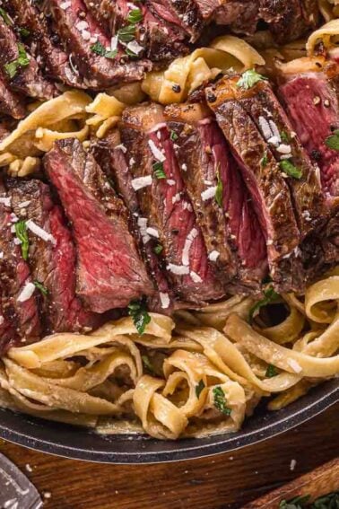 A skillet filled with sliced medium-rare steak served on top of fettuccine alfredo pasta with a creamy sauce, garnished with herbs and grated cheese.