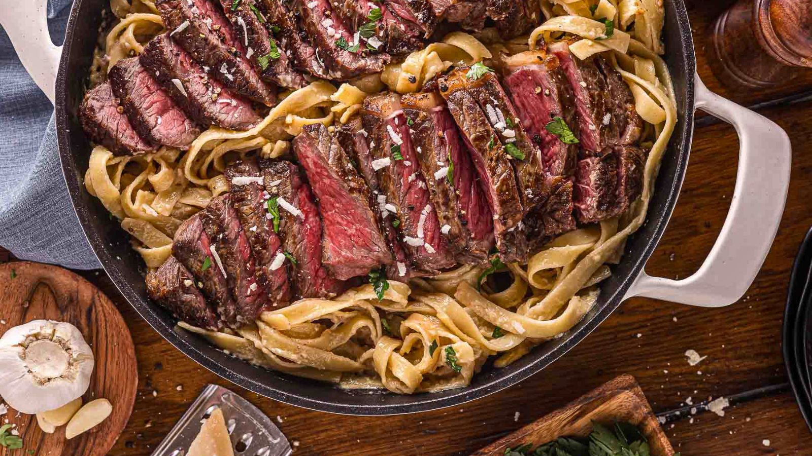 A skillet filled with sliced medium-rare steak served on top of fettuccine alfredo pasta with a creamy sauce, garnished with herbs and grated cheese.
