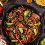 A skillet of cooked steak strips with sautéed bell peppers and onions, surrounded by toppings like avocado, sour cream, salsa, tomatoes, and tortillas.