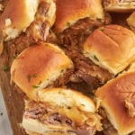 A close-up of several french dip sliders with beef and onion on a wooden cutting board, topped with chopped herbs.
