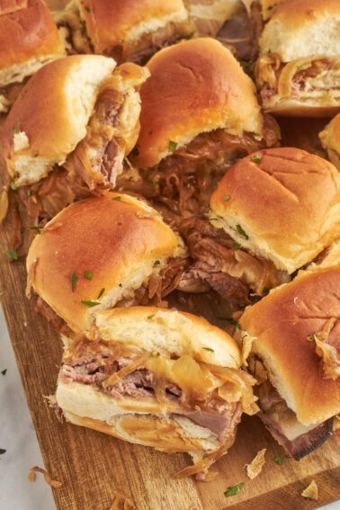 A close-up of several french dip sliders with beef and onion on a wooden cutting board, topped with chopped herbs.