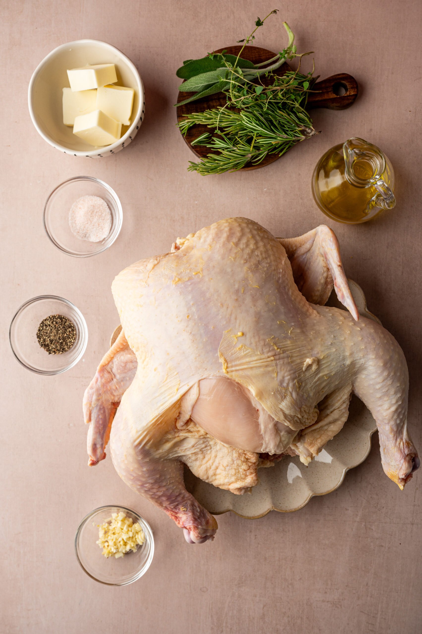 Raw whole chicken on a plate surrounded by bowls of butter, salt, pepper, minced garlic, olive oil, and fresh herbs on a beige surface.