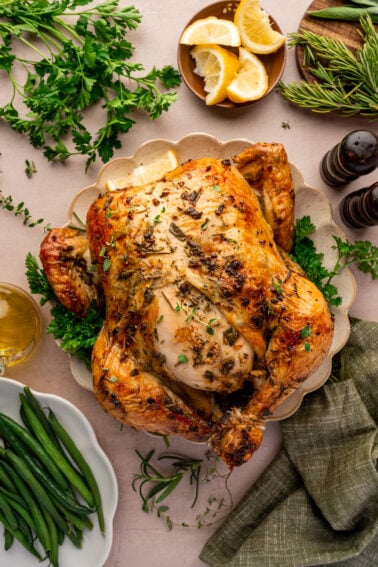 Roast chicken garnished with herbs on a platter, surrounded by lemon wedges, green beans, parsley, rosemary, salt and pepper shakers, and a green napkin.