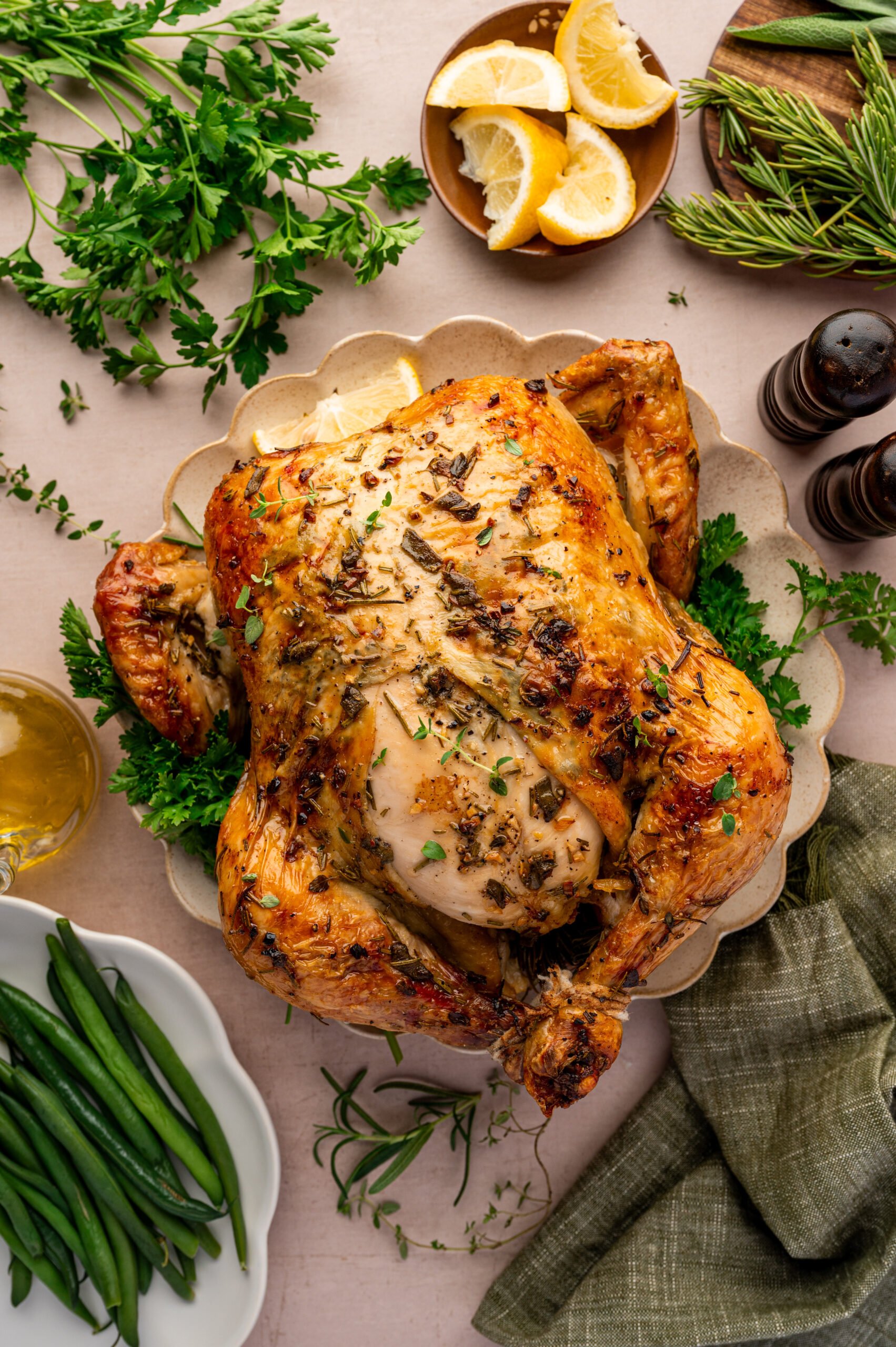 Roast chicken garnished with herbs on a platter, surrounded by lemon wedges, green beans, parsley, rosemary, salt and pepper shakers, and a green napkin.
