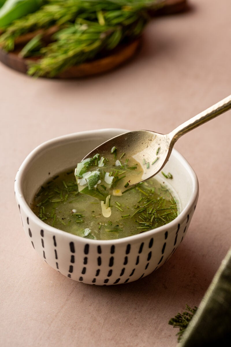 A spoon lifts herbs and butter from a small bowl of clear broth, with fresh herbs in the background.