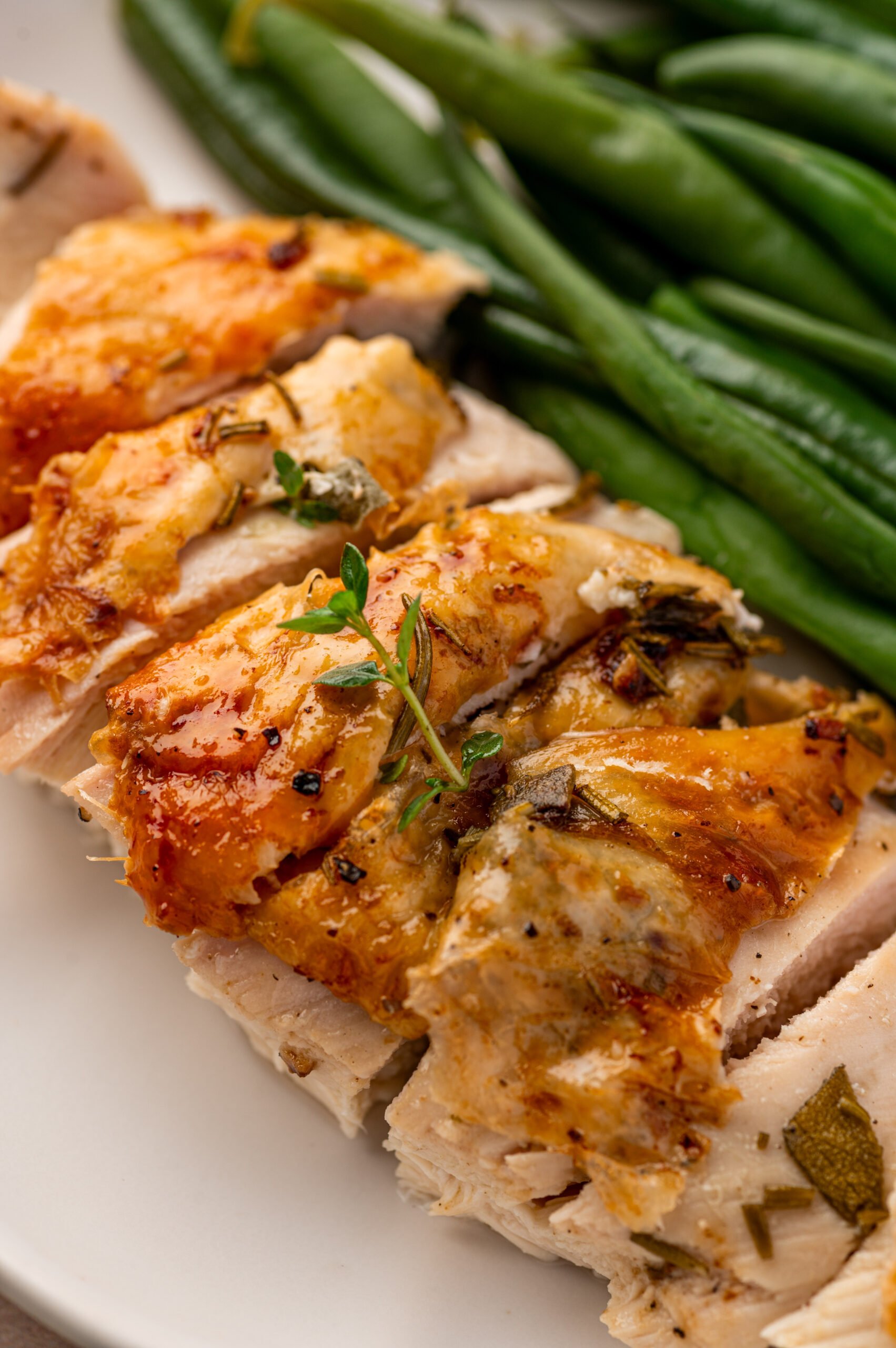 Sliced herb roasted chicken breast garnished with herbs, served next to steamed green beans on a white plate.