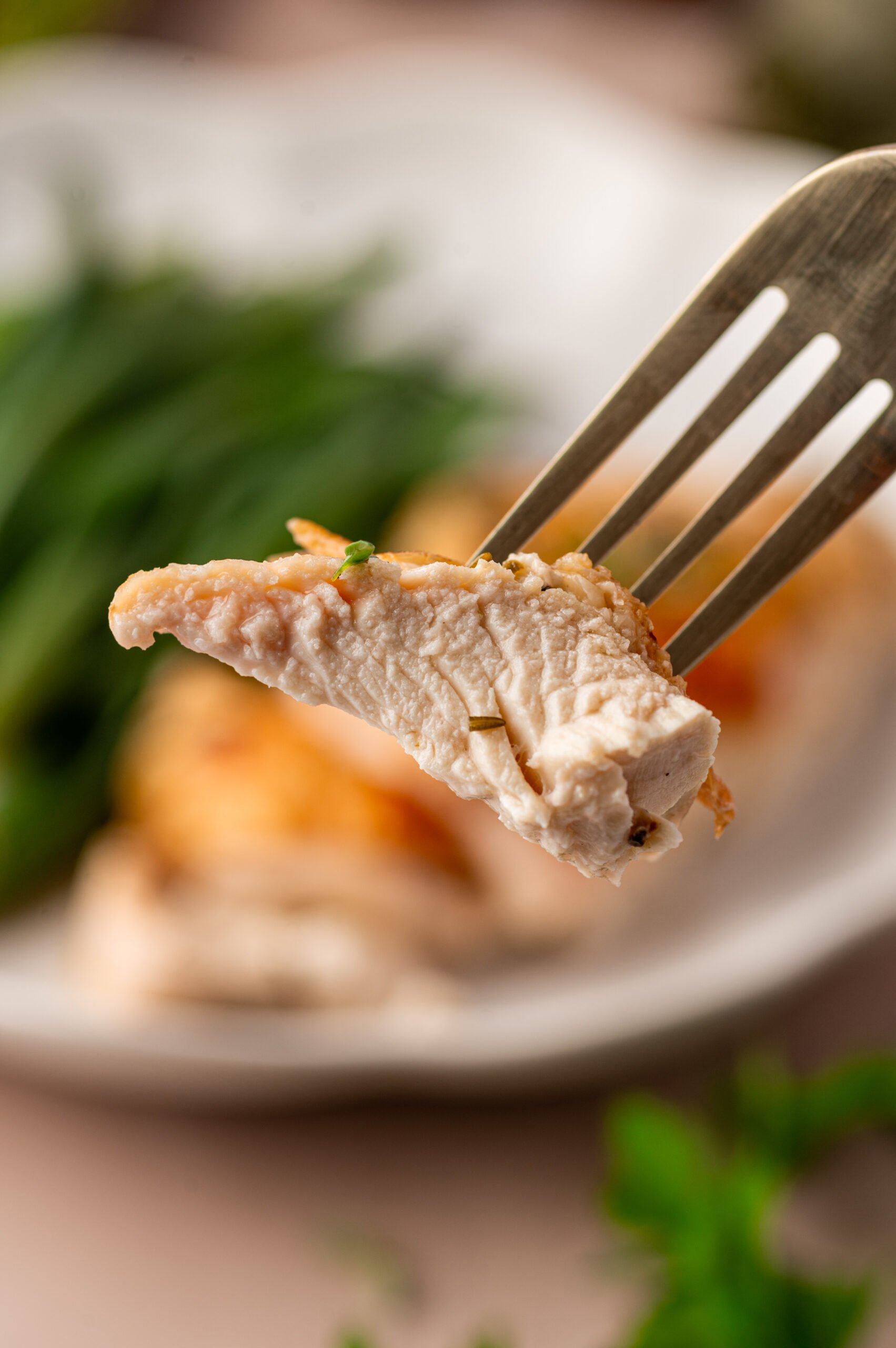 A fork holds a slice of cooked chicken breast, with fresh herbs visible; a plate with more chicken and greens is blurred in the background.