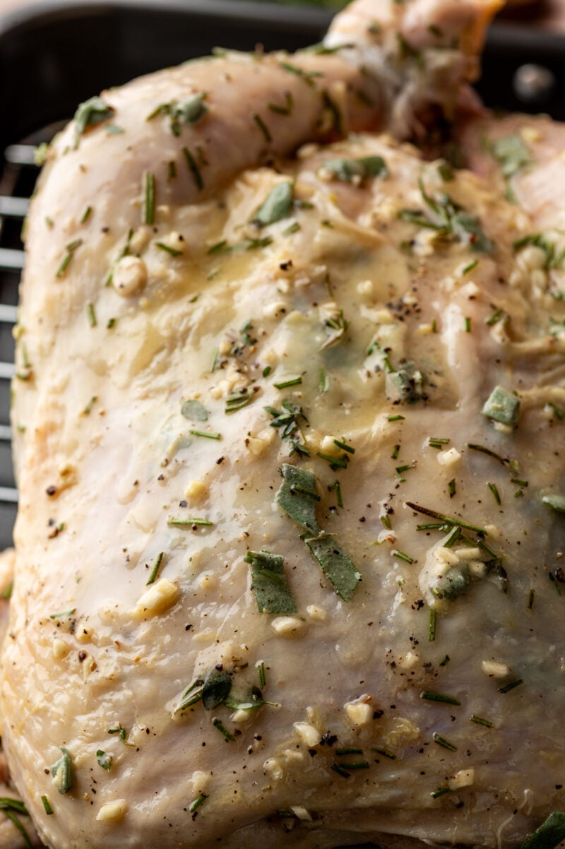 Close-up of raw chicken coated with chopped herbs, minced garlic, and black pepper, placed on a roasting rack.