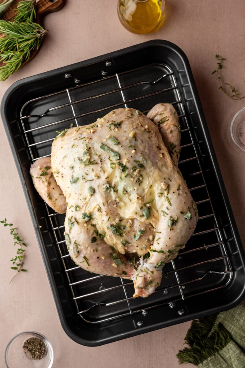 Raw whole chicken coated with herbs and butter placed on a metal rack inside a roasting pan, surrounded by small bowls of spices, herbs, and oil.