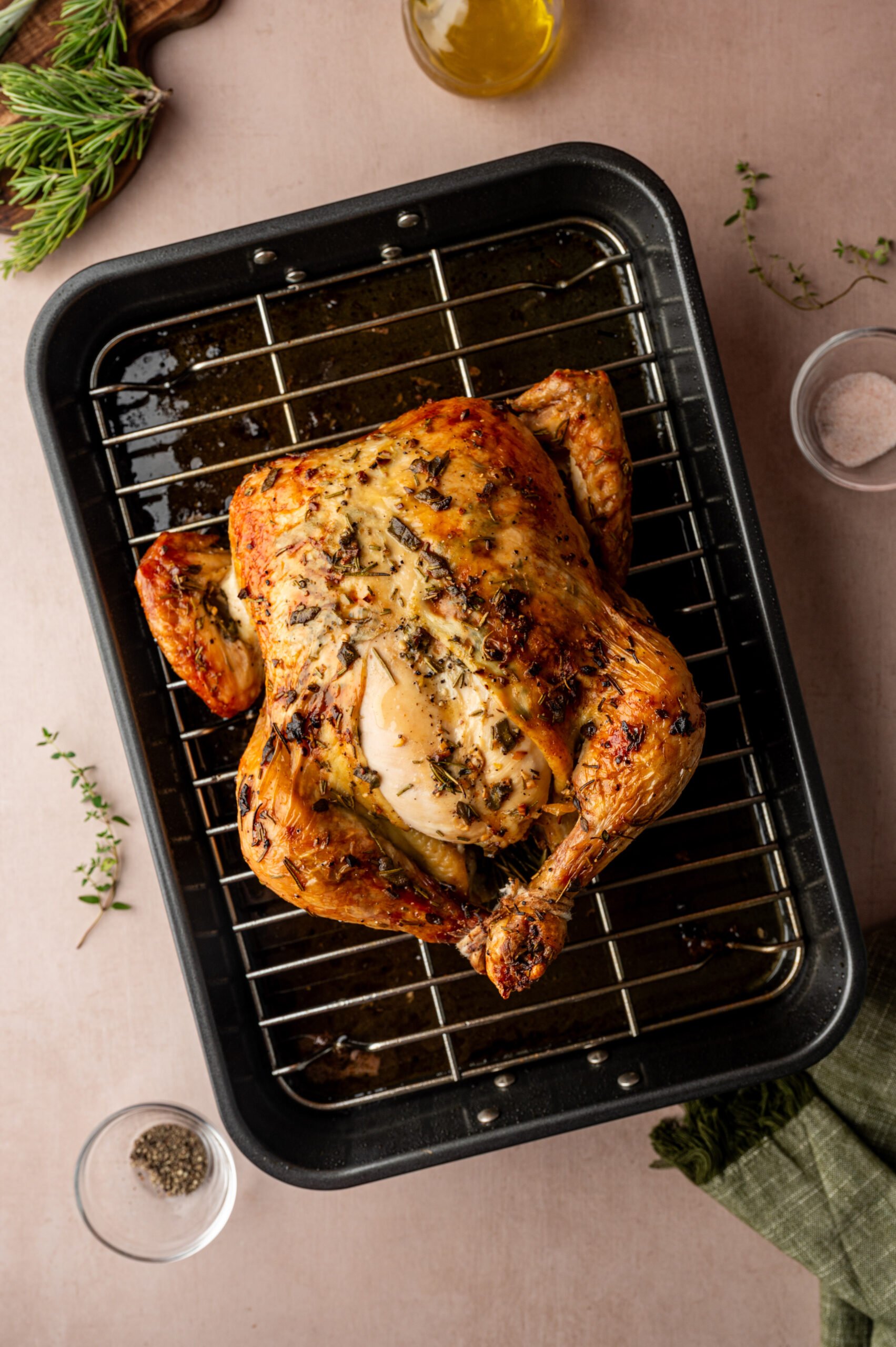 A whole roasted chicken with herbs sits on a rack in a roasting pan, surrounded by small bowls of seasoning on a light-colored surface.