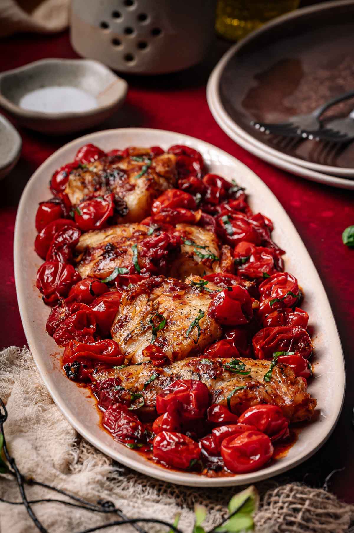 Pan seared cod served on an oval plate with roasted cherry tomatoes and fresh herbs, set on a red tablecloth with dishes and utensils in the background.