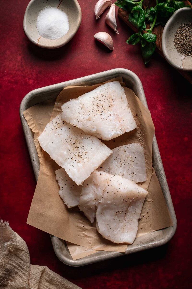 Raw white cod fillets on parchment paper in a metal tray, seasoned with pepper, surrounded by garlic, basil, salt, and pepper on a red surface.