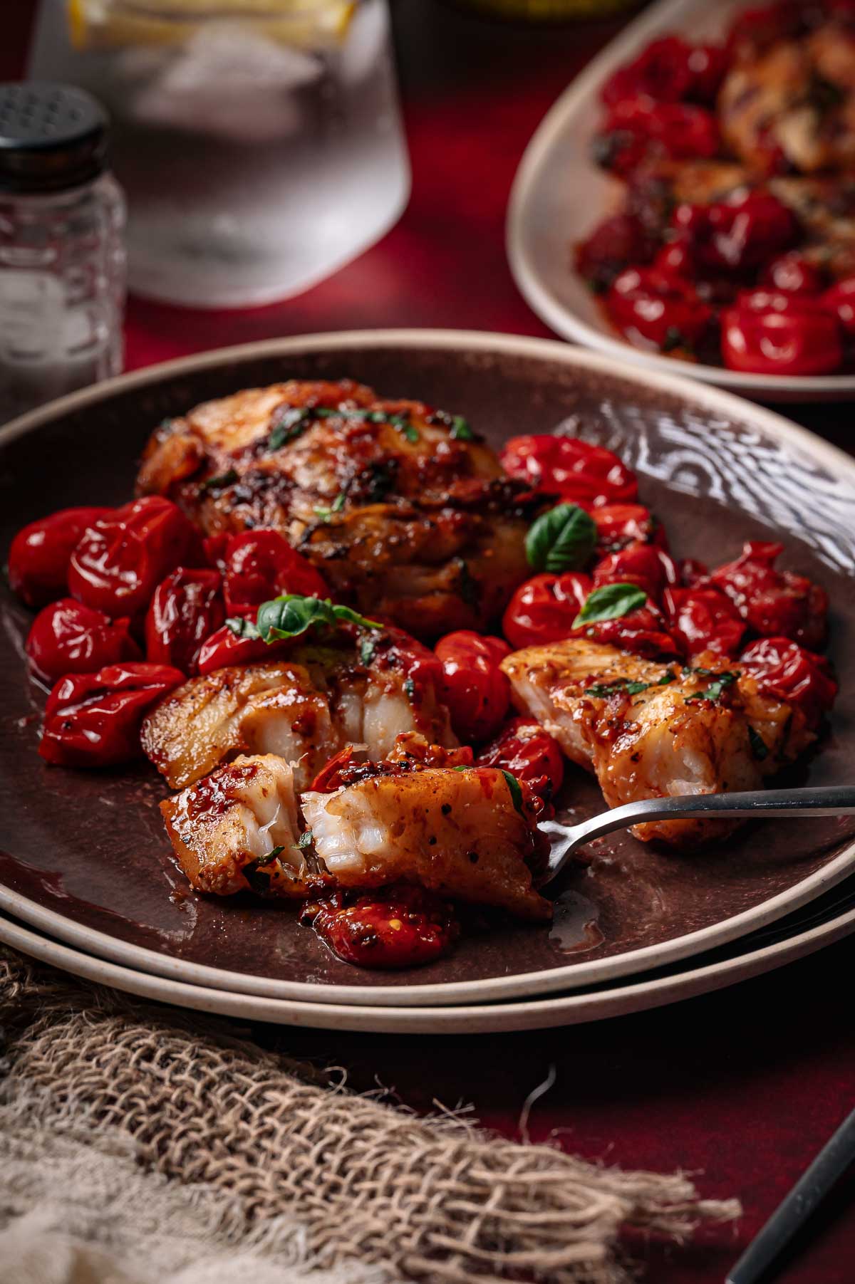 A plate of pan seared white fish garnished with herbs, served with blistered cherry tomatoes and a fork, on a rustic brown dish.