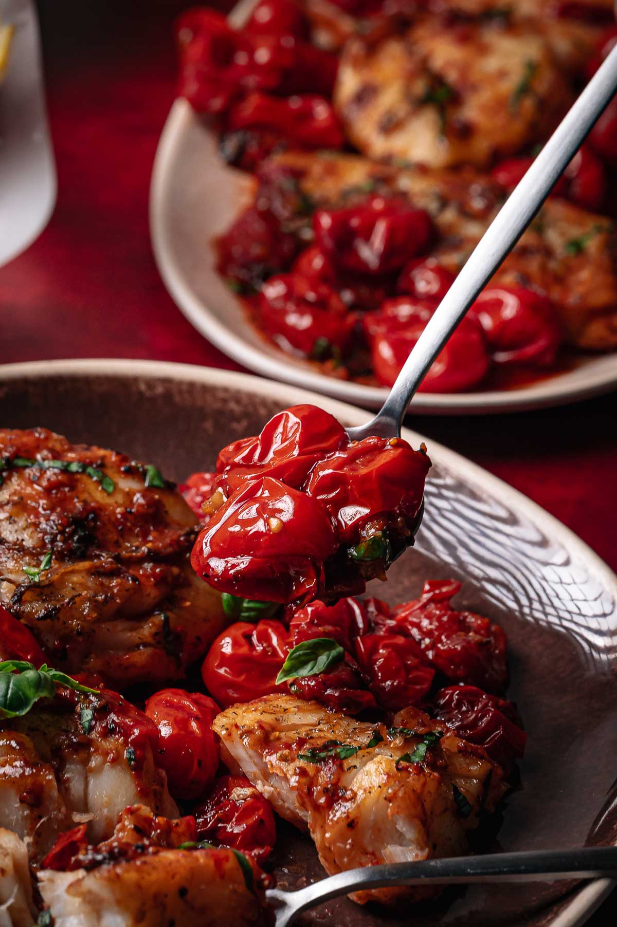 A close-up of a fork holding roasted cherry tomatoes above a plate of seared cod garnished with herbs, with another plate of the same dish in the background.