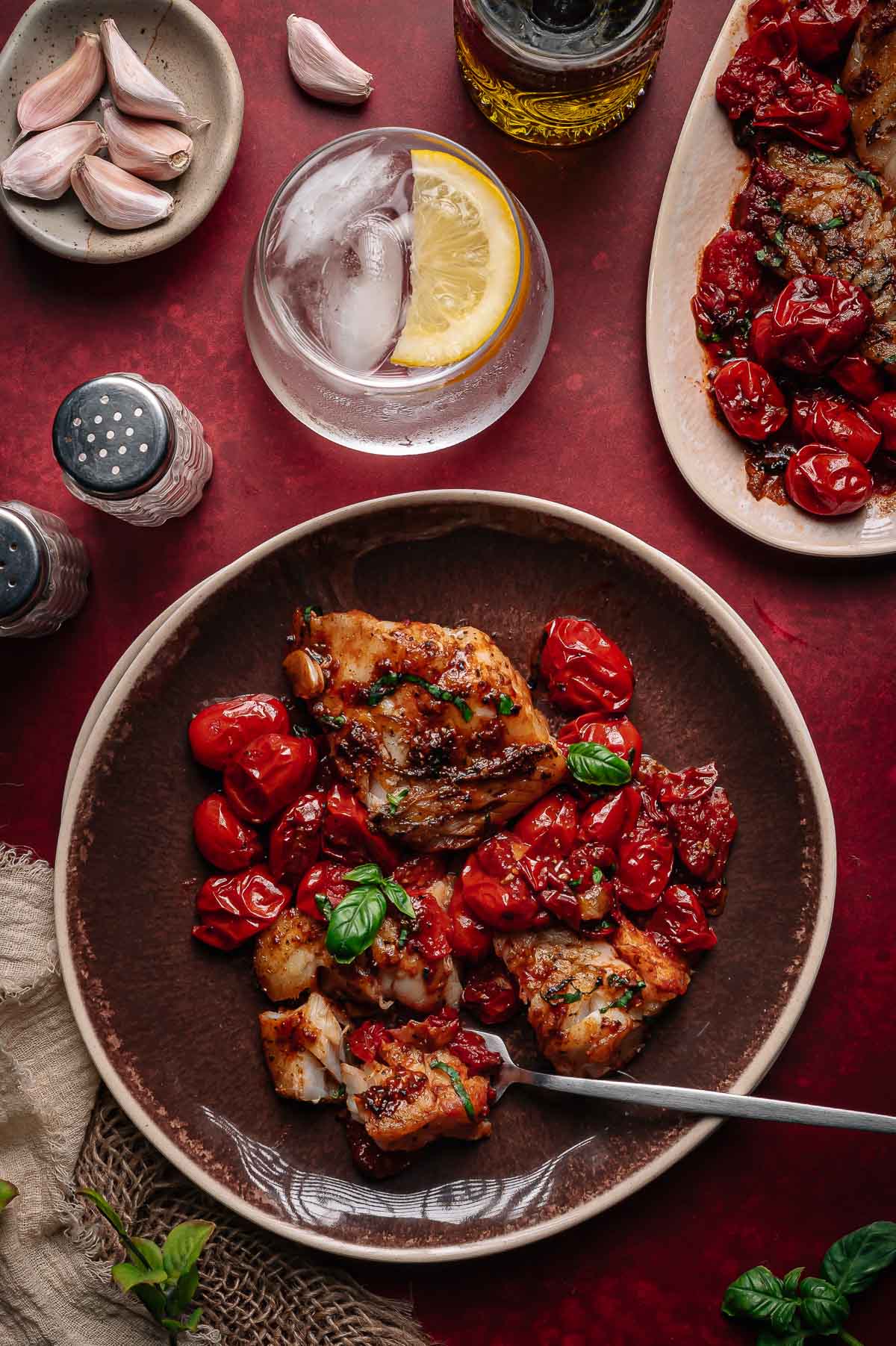 A brown plate with cooked cod, roasted cherry tomatoes, and fresh basil, next to a drink with ice and lemon, garlic cloves, and seasonings on a red table.