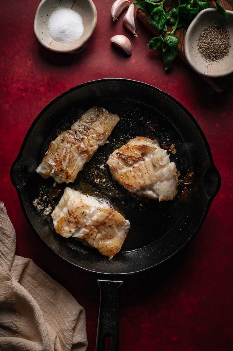 Three pieces of cooked white cod in a cast iron skillet, with salt, pepper, garlic, and herbs on a red surface.