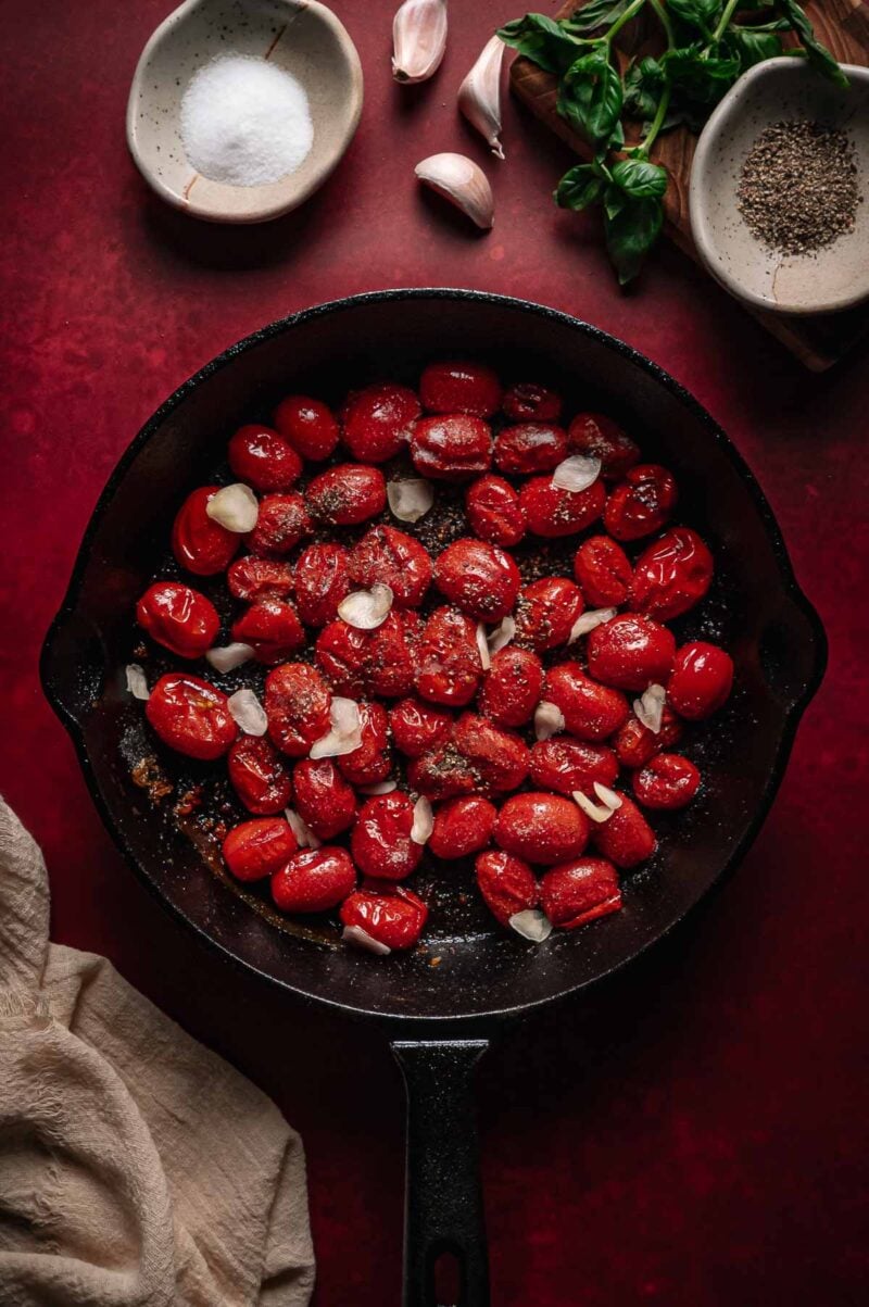 A cast iron skillet filled with cherry tomatoes, sliced garlic, and seasonings; surrounded by bowls of salt, pepper, garlic cloves, basil, and a beige cloth on a red surface.
