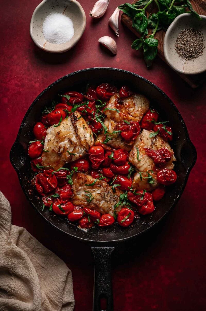 A cast iron skillet with cooked cod, cherry tomatoes, and chopped herbs, surrounded by bowls of salt, pepper, garlic cloves, and basil on a red surface.