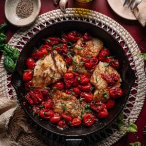 A cast iron skillet with cooked cod fillets and roasted cherry tomatoes, garnished with herbs, surrounded by garlic, olive oil, and seasonings on a red surface.