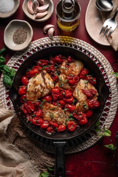 A cast iron skillet with cooked cod fillets and roasted cherry tomatoes, garnished with herbs, surrounded by garlic, olive oil, and seasonings on a red surface.