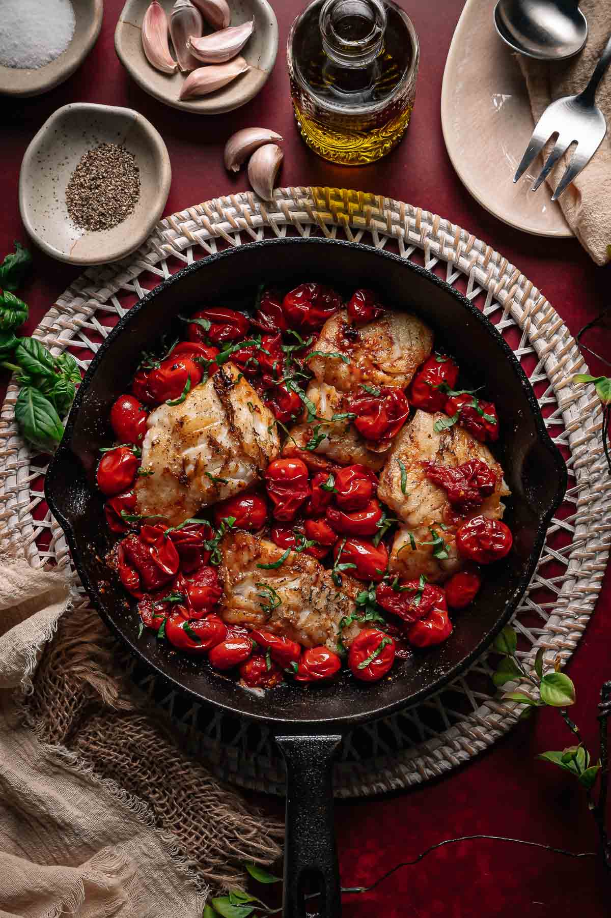 A cast iron skillet with cooked cod fillets and roasted cherry tomatoes, garnished with herbs, surrounded by garlic, olive oil, and seasonings on a red surface.