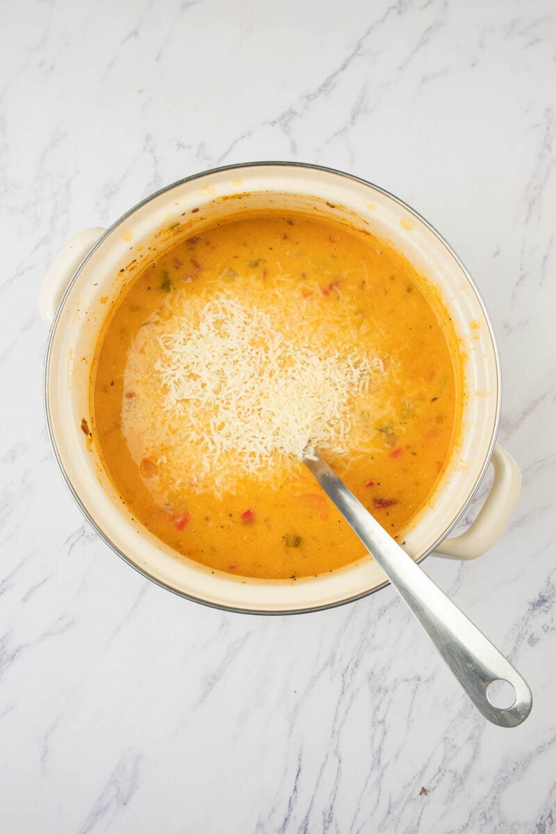 A pot of creamy soup topped with shredded cheese sits on a white marble surface, with a metal ladle resting inside.
