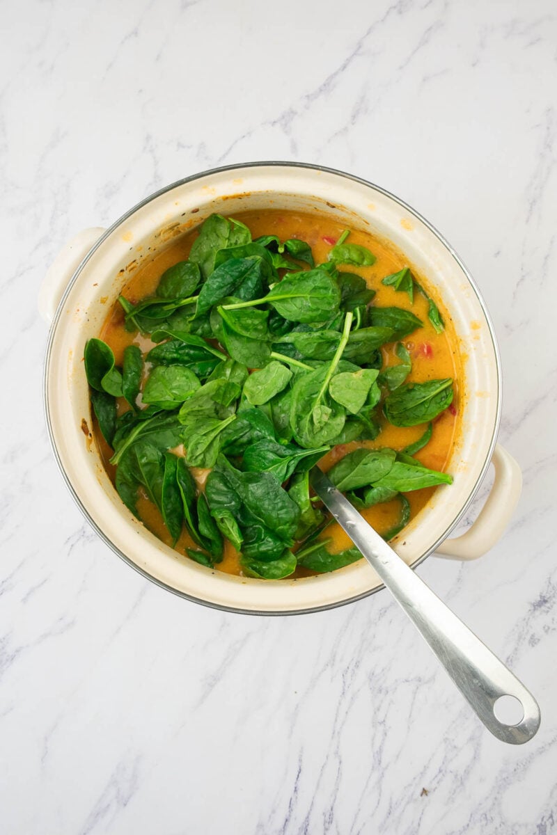 A pot of soup with fresh spinach leaves on top being wilted in and a metal ladle, placed on a white marble surface.
