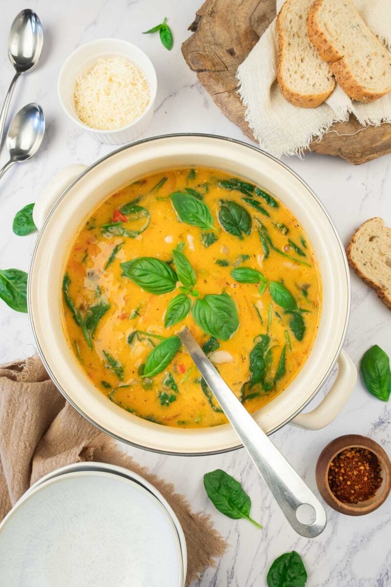 A pot of creamy white bean soup with chicken and basil leaves, surrounded by sliced bread, grated cheese, red pepper flakes, and spoons on a marble surface.