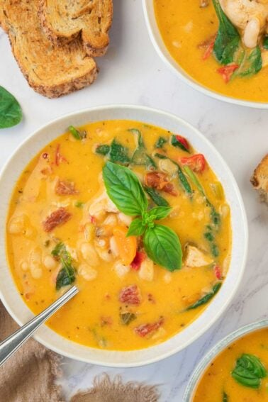 A bowl of creamy soup with white beans, spinach, chicken, and herbs, garnished with basil, next to slices of toasted bread.