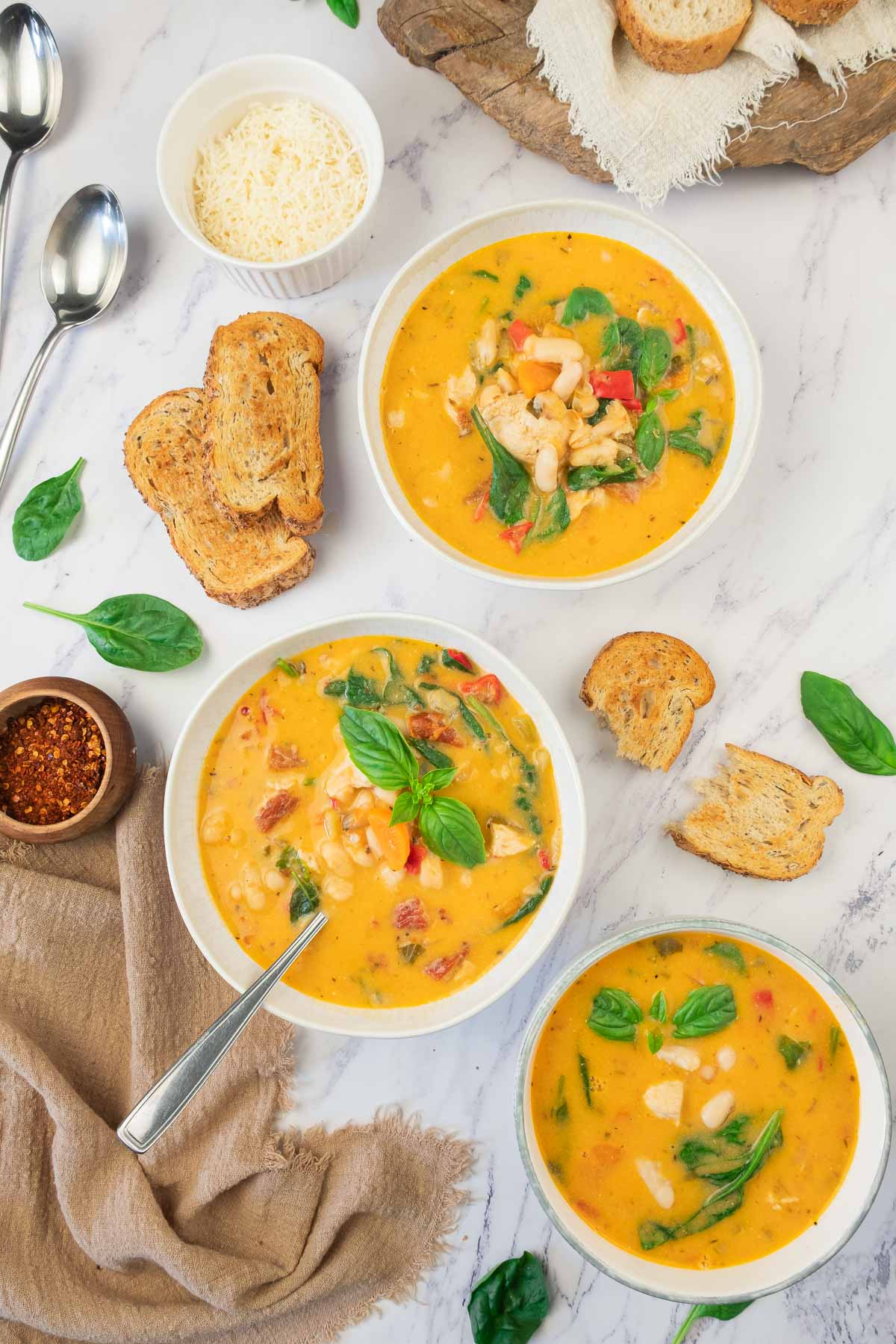 Three bowls of creamy tuscan white bean soup with white chicken, spinach, and tomatoes, surrounded by toasted bread, grated cheese, dried chili flakes, spoons, and fresh basil leaves on a white marble surface.