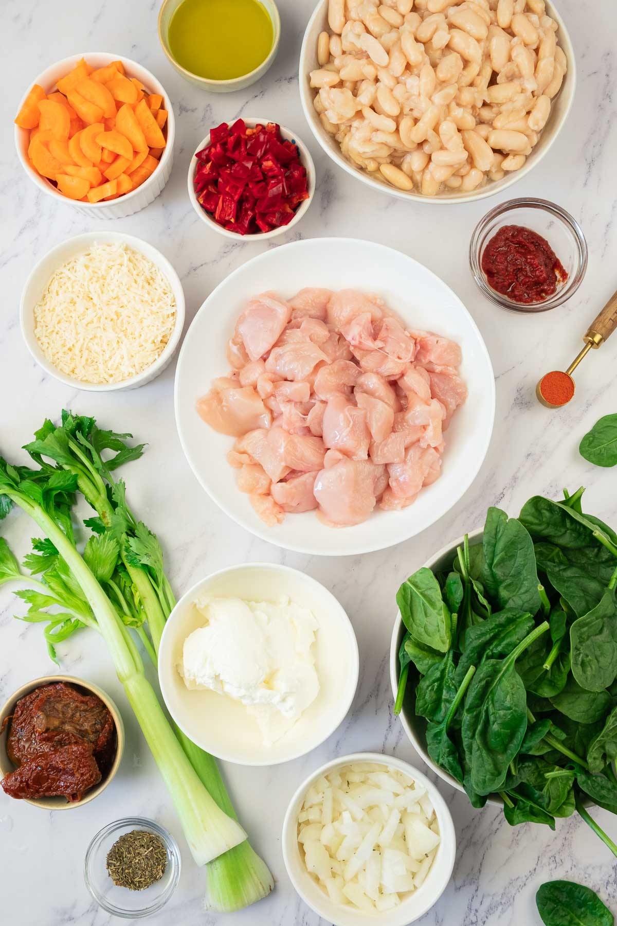 A variety of ingredients arranged on a white surface, including raw chicken, spinach, white beans, carrots, celery, onion, cheese, ricotta, spices, and olive oil.