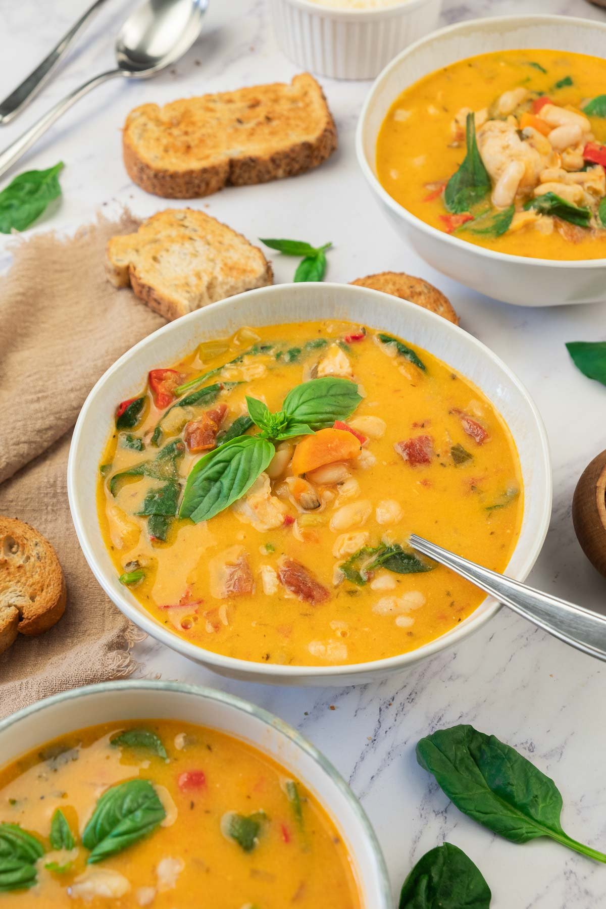 Three bowls of creamy soup with white beans, tomatoes, spinach, and fresh basil, accompanied by toasted bread slices on a marble surface.