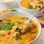 A spoonful of creamy tuscan white bean soup with chicken is held above a bowl, with another bowl of soup, bread, and a small dish in the background.