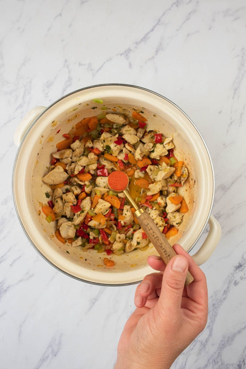 A hand holds a measuring spoon with paprika over a pot of chopped chicken and vegetables on a marble countertop.