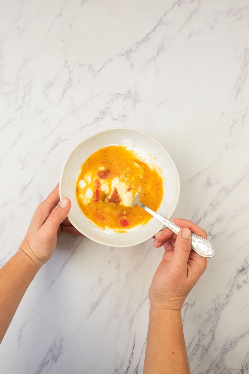 A person holds a white bowl showing sour cream being tempered with a little bit of broth and a fork over a white marble surface.