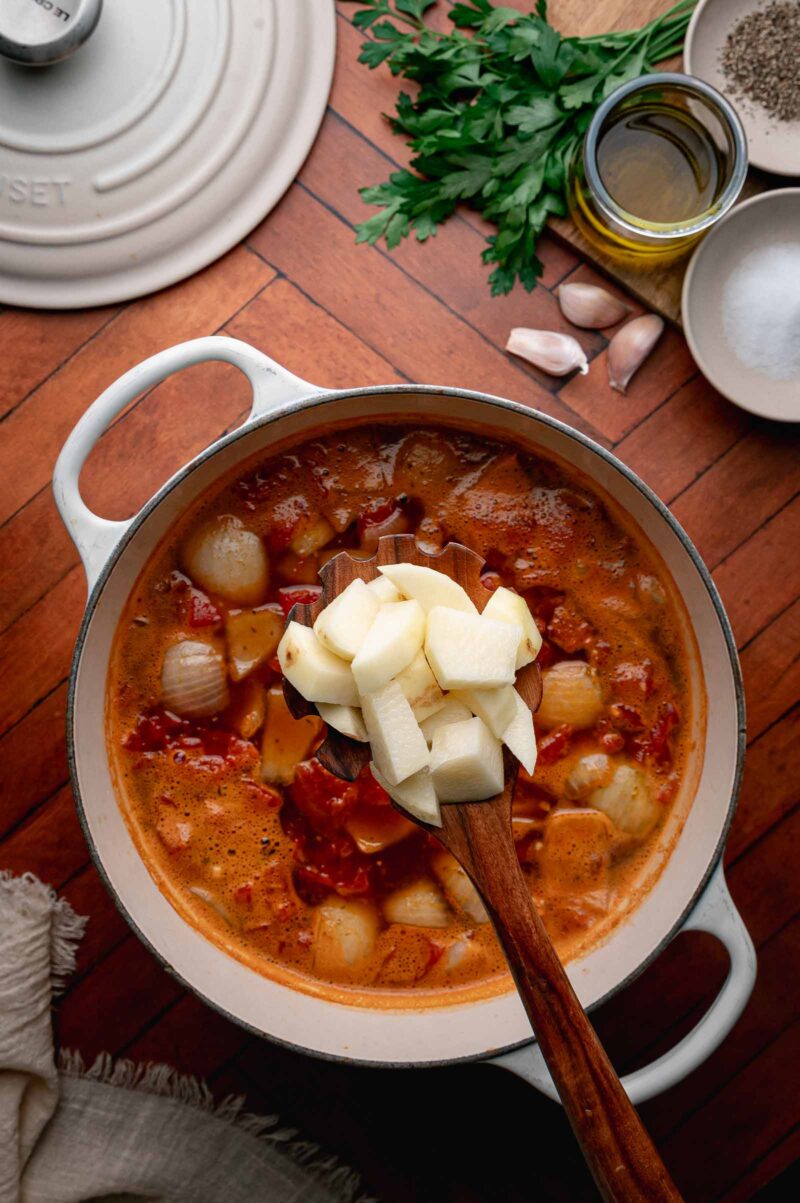 A wooden spoon holds chopped potatoes over a pot of soup with onions and tomatoes on a wooden table, surrounded by herbs, garlic, oil, salt, and pepper.