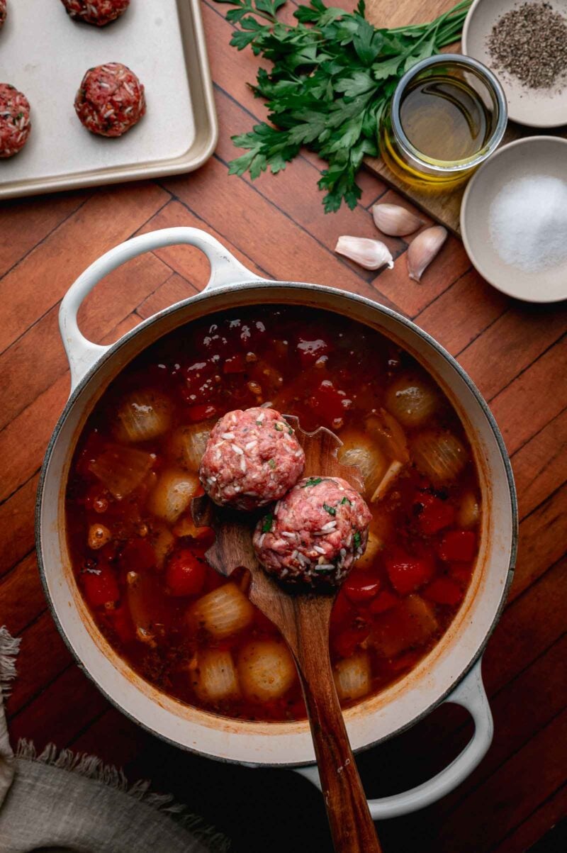 A pot of albondigas soup with onions sits on a wooden surface. A wooden spoon holds two raw meatballs above the pot. Ingredients like parsley, garlic, olive oil, and seasonings are nearby.