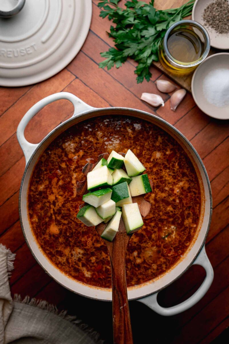 A pot of albondigas soup on a stove, with a wooden spoon holding chopped zucchini above the pot. Ingredients like parsley, garlic, and olive oil are on the counter nearby.