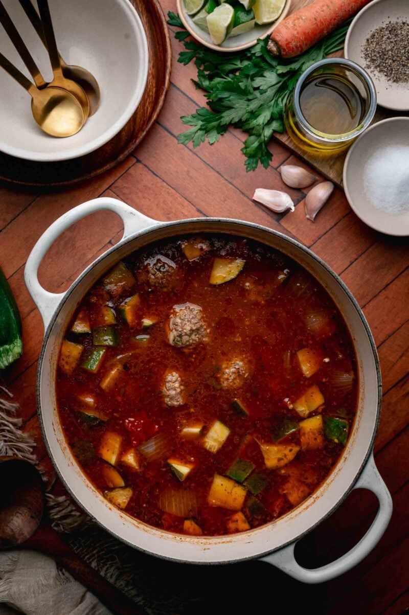 A pot of albondigas soup sits on a wooden table, surrounded by bowls, parsley, a carrot, lime wedges, garlic, oil, and seasonings.