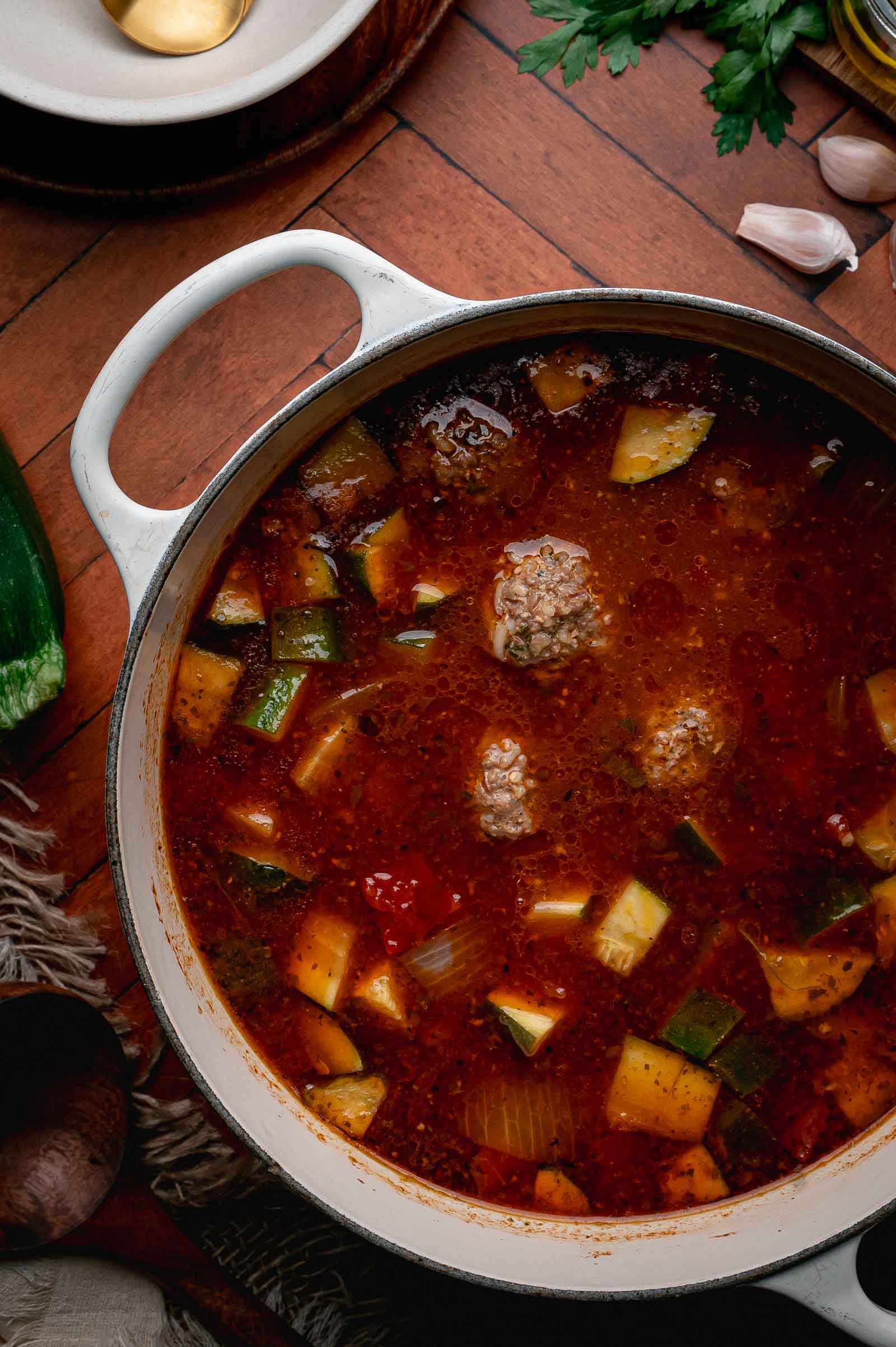 A pot of albondigas soupwith meatballs, tomatoes, zucchini, and onions in a red broth on a wooden surface, surrounded by herbs and kitchen items.