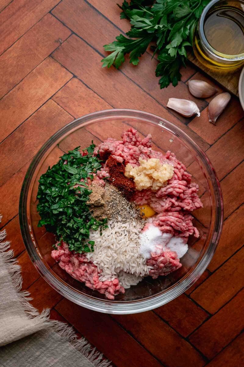 A glass bowl containing ground meat, chopped parsley, uncooked rice, spices, salt, a raw egg, and minced garlic, set on a wooden surface with herbs, oil, and garlic nearby.